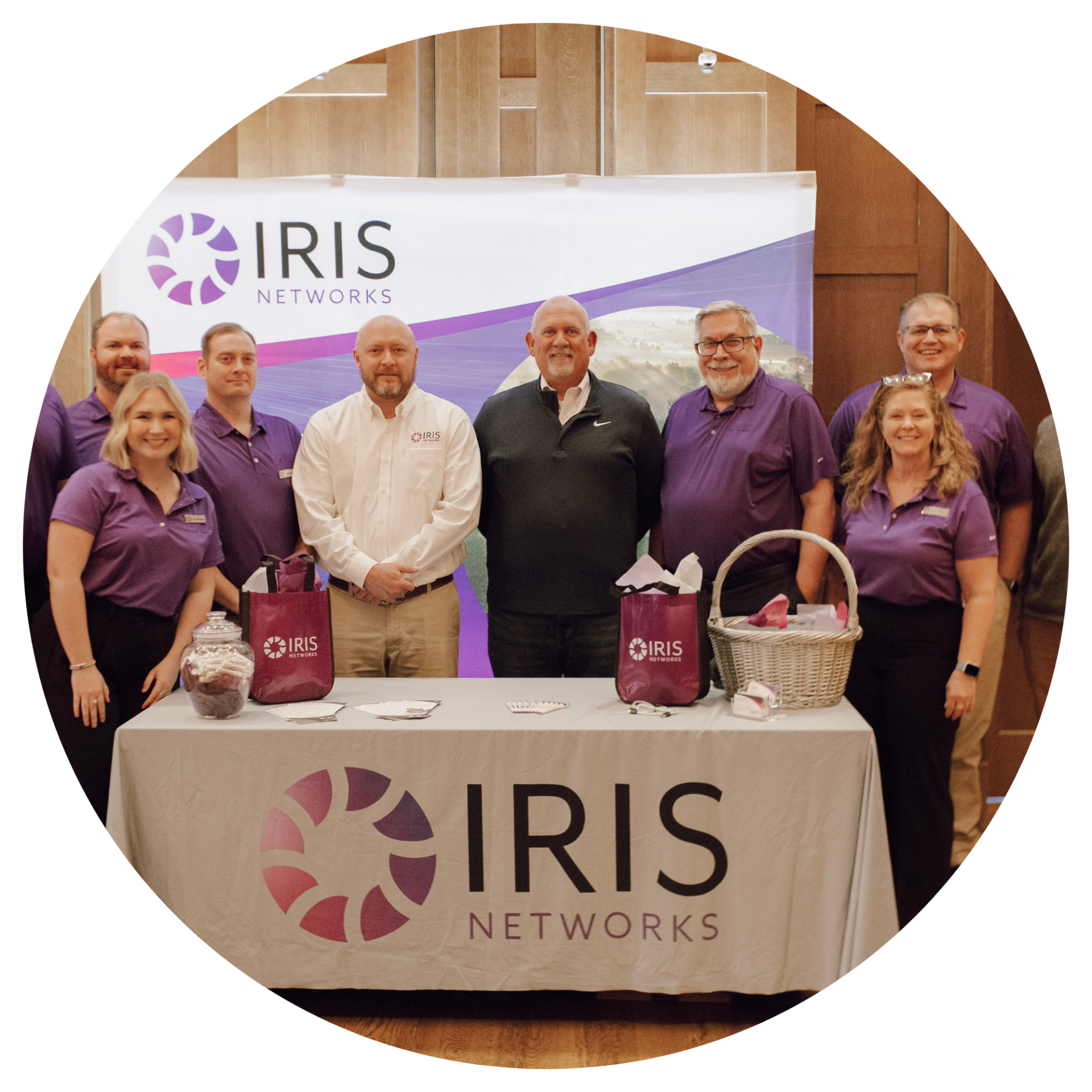 IRIS Networks team wearing purple white and black, stand by a table with IRIS Networks display items.